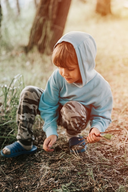 Schattige kleine vijfjarige jongen in blauwe hoodie verzamelt takjes kreupelhout voor kampvuur in het dorp op de natuur buiten op een zomeravond. authentieke platteland en het rustieke leven van kinderen. gloed