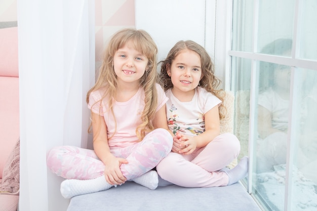 Schattige kleine zusjes zittend op de vensterbank thuis, portret van mooie kleine meisjes op kamer vensterbank in de winter.