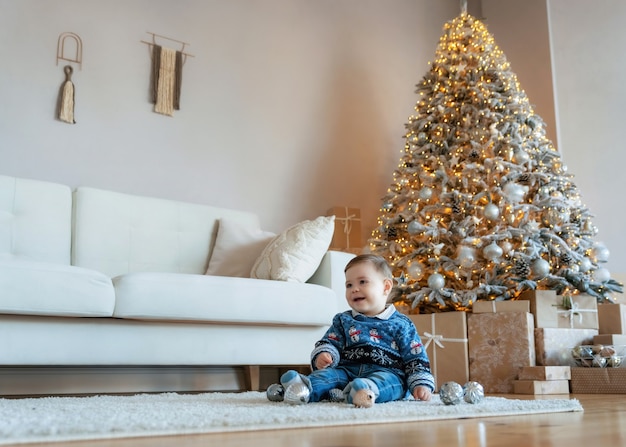 Schattige lachende babyjongen zittend op een wit wollen tapijt in een lichte, gezellige woonkamer met een kerstboom en veel geschenkdozen. eerste kerst. oudjaarsavond.