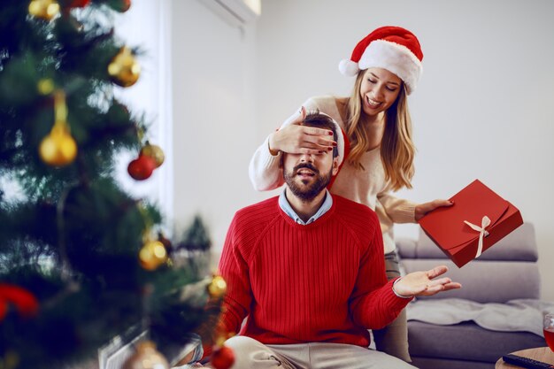 Schattige lachende blanke vrouw met cadeau en voor de ogen van haar vriendje