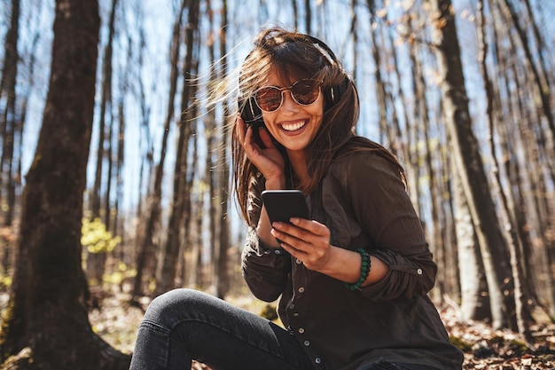 Schattige lachende jonge vrouw, met smartphone in haar hand en koptelefoon op haar hoofd, zit op de herfstbladeren in de vroege herfst zonnige dag en luistert naar muziek.