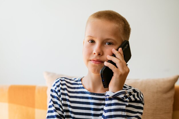 Schattige lachende kleine jongen zittend op de bank en praten over de telefoon thuis in casual kleding op een zon...