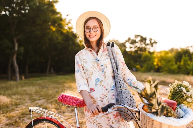 Schattige lachende meisje in bril op fiets met mand vol fruit en wilde bloemen gelukkig in de camera kijken in park