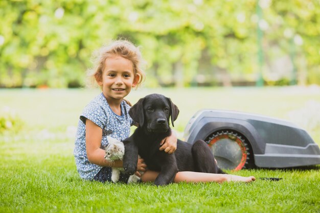 Schattige lachende meisje zittend op vers gemaaid gazon omarmen haar huisdieren zwarte puppy en witte pluizige kitten Kinderen en huisdieren brengen graag tijd door op vers gras De gazonrobot op de achtergrond