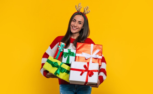 Schattige lachende opgewonden jonge brunette vrouw in herten hoed, met veel kleurrijke kerst geschenkdozen in handen geïsoleerd op gele achtergrond.