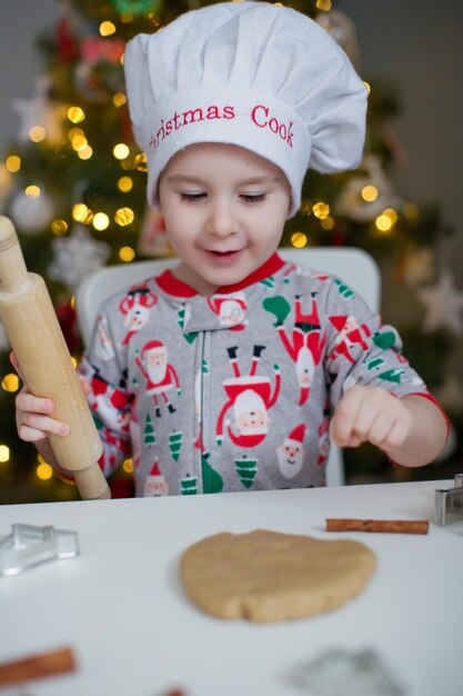 Schattige peuterjongen die kerstkoekjes maakt op een witte tafel in de buurt van de kerstboom met lichtjes Kerstkookconcept