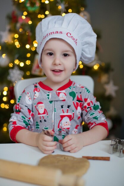 Schattige peuterjongen die kerstkoekjes maakt op een witte tafel in de buurt van de kerstboom met lichtjes Kerstkookconcept