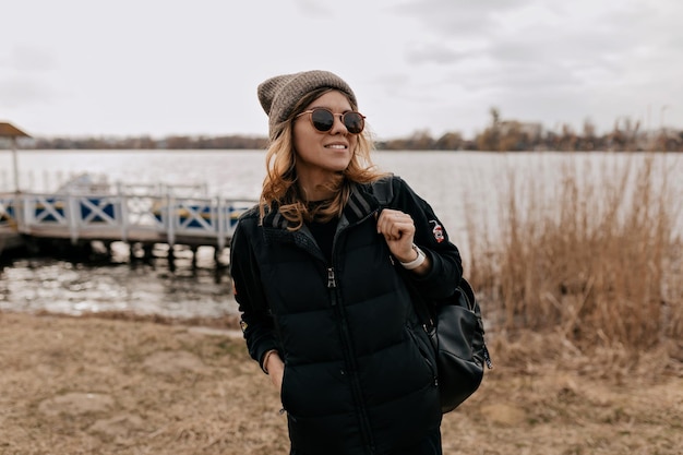 Schattige schattige dame in stijlvolle lente-outfit kijkt weg met een gelukkige glimlach tijdens het wandelen op het strand in de lente