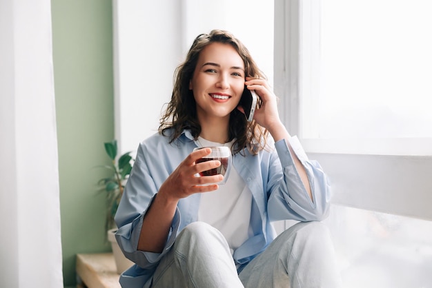 Schattige schattige grappige vrouw met een mok zwarte thee praten aan de telefoon zittend op de vensterbank in de keuken plezier maken en genieten van een aangenaam gesprek met haar zus of beste vriendin