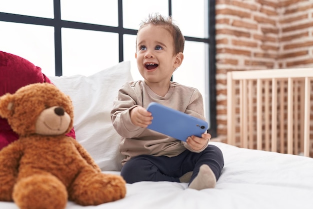 Schattige Spaanse jongen met smartphone zittend op bed in de slaapkamer