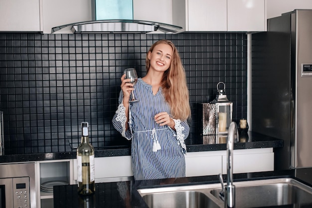 Schattige vrolijke jongedame in een gestreepte jurk poseert in de keuken met een glas wijn in haar hand