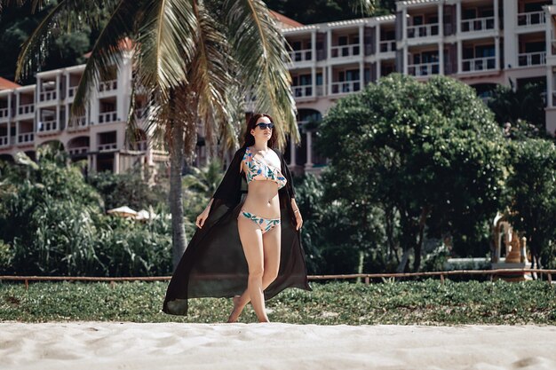 Schattige zelfverzekerde vrouw in een bikini en een lang zwart gewaad wandelen langs het zandstrand tegen de achtergrond van huizen en palmbomen. Concept van ontspanning