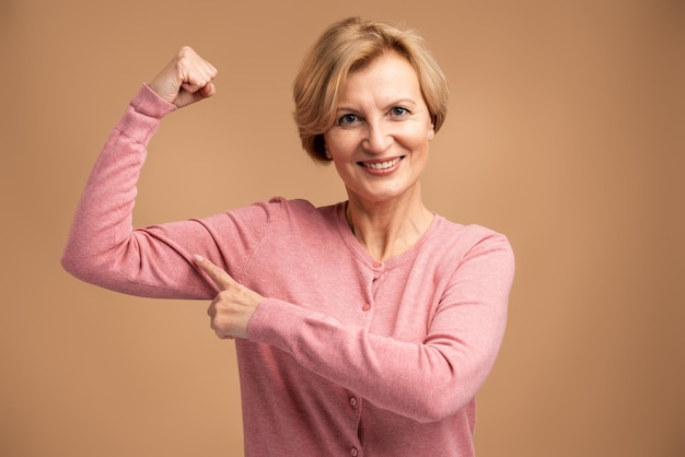 Schattige zelfverzekerde vrouw wijzende vinger op haar armspier, pompen in de sportschool, korting. Indoor studio-opname geïsoleerd op beige background