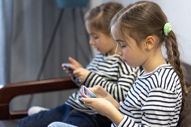 Schattige zusjes gebruiken telefoons terwijl ze thuis op de bank zitten.