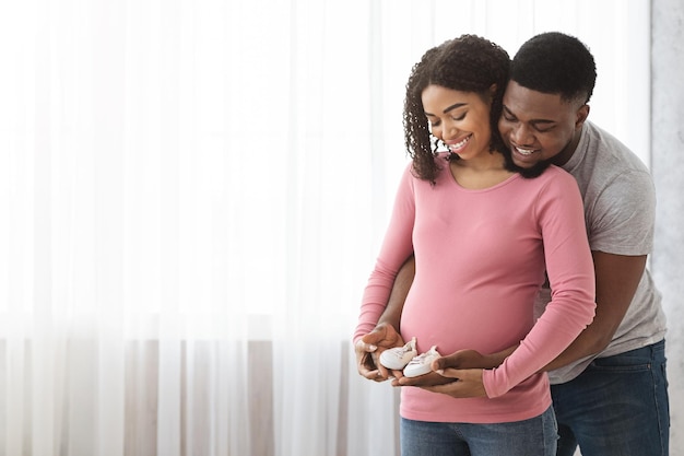 Schattige zwarte verwachtende familie die kleine babyschoentjes bij elkaar houdt, thuis naast het raam staat, vrije ruimte