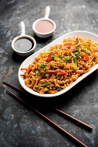 Schezwan veg noodles is a spicy and tasty stir fried flat Hakka noodles with sauce and veggies. served with chopsticks. selective focus