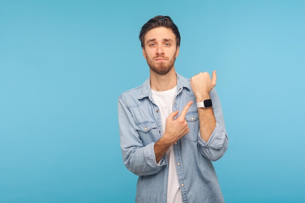 Schiet op. Portret van een punctuele man in een spijkerhemd van een werknemer, wijzend op een mock-up van een polshorloge en uiting gevend aan ontevredenheid over de late tijd, deadline. indoor studio-opname geïsoleerd op blauwe achtergrond