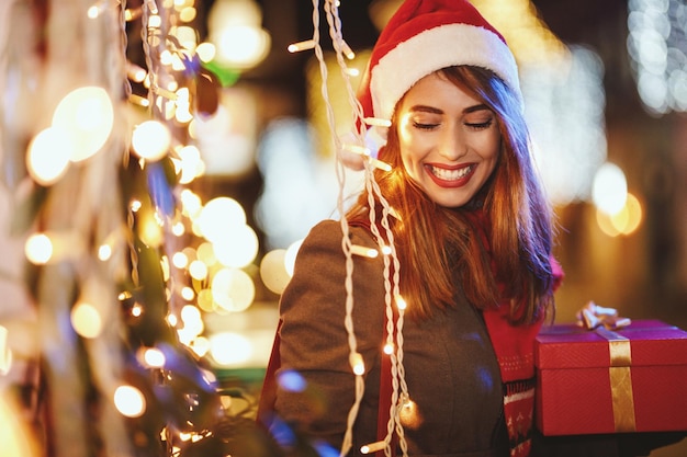 Schieten van een vrolijke jonge vrouw met een rode huidige doos heeft plezier in de stadsstraat met kerst.