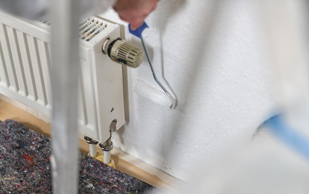 schilder die met verfroller werkt om achter een radiator de muur met witte kleur te schilderen. doe het zelf concept afbeelding