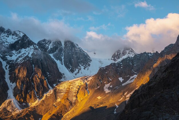 Schilderachtig berglandschap met besneeuwde bergtop en gletsjer in lage wolken in gouden zonsopgangkleuren Kleurrijk uitzicht op de bergen naar verticale gletsjer met ijsval in gouden ochtendzonlicht in lage wolken