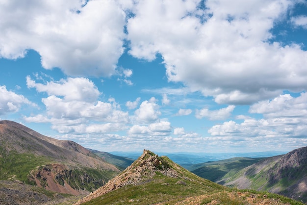 Schilderachtig groen landschap met zonovergoten puntige stenen heuvel met grassen en bloemen onder wolken in de blauwe lucht Kleurrijk alpenlandschap met scherpe rotsen met gras en bloemen in zonlicht bij wisselvallig weer