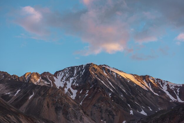 Schilderachtig landschap met hoge sneeuwberg met scherpe rotsachtige top in gouden zonlicht onder wolken van zonsondergangskleur bij veranderlijk weer Kleurrijk uitzicht op grote bergtop onder bewolkte zonsondergangshemel