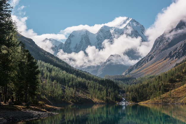 Schilderachtig landschap met turkoois bergmeer in de herfstvallei tegen grote sneeuwbergen in lage wolken in de ochtendzon Alpenmeer met uitzicht op de zonovergoten hoge besneeuwde bergketen in lage bewolking