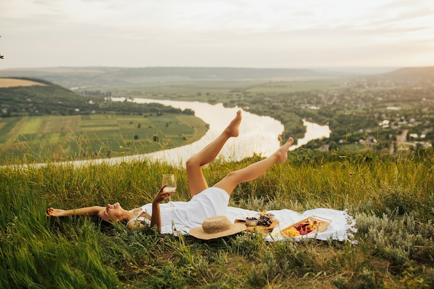 Schitterend jong meisje in een witte zomerjurk die op gras ligt en een picknick op een schilderachtige plek heeft.