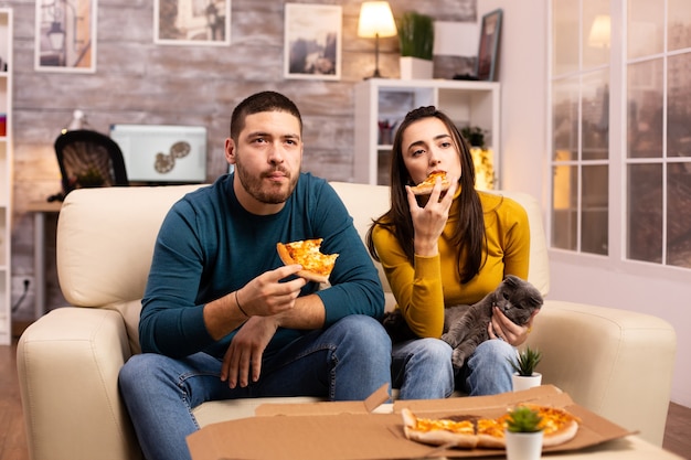 Schitterend jong stel dat pizza eet terwijl ze tv kijken in de woonkamer op de bank