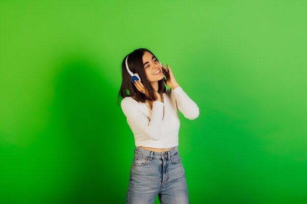 Schitterend lachend meisje in witte blouse en spijkerbroek luisteren muziek in koptelefoon terwijl staande op groen oppervlak.