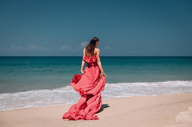 Schitterend meisje in het zonnige exotische strand aan de oceaan. De jonge vrouw draagt een verbazingwekkende rode jurk die wappert in de lichte bries, achteraanzicht; mode-concept.