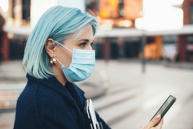 Schitterende blanke ondernemer met blauw haar poseren met moderne gadgets terwijl ze een anti-griepmasker draagt