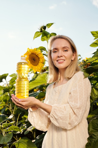 Schitterende blonde jonge vrouw die een fles zonnebloemolie vasthoudt terwijl ze tegen grote bloemen staat voor de camera in het veld