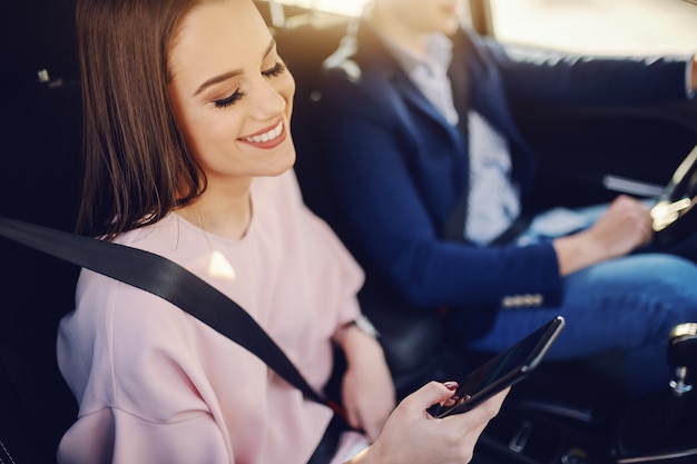 Schitterende brunette met brede glimlach, gekleed elegante zitten in de auto en het gebruik van slimme telefoon terwijl haar vriendje in de achtergrond rijden.