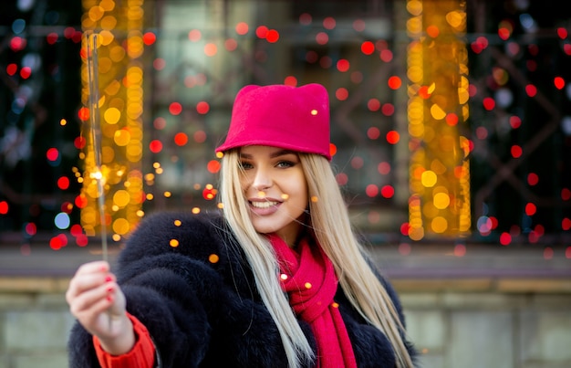 Schitterende glimlachende blonde vrouw die winterkleding draagt, die Kerstmis viert bij de slingers vage achtergrond. Lege ruimte