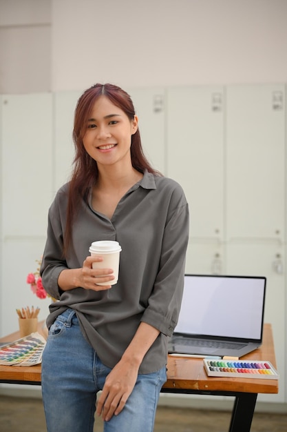Schitterende jonge Aziatische vrouwelijke grafisch ontwerper die op tafel leunt en een kopje koffie drinkt