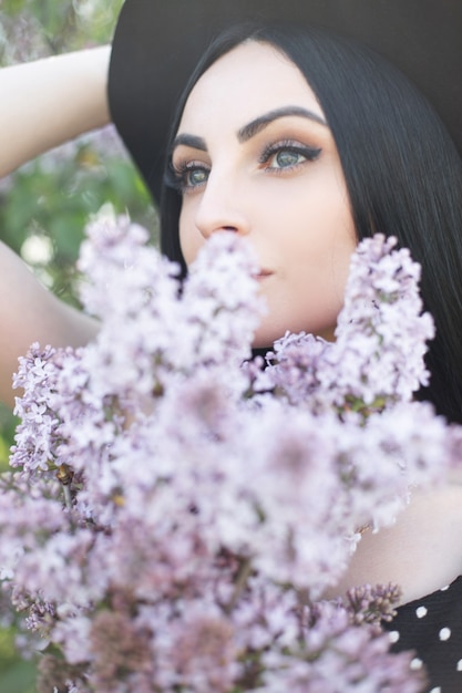 Schitterende jonge vrouw met verbazende ogen met schone huid en lila boeketbloemen op aard. Aantrekkelijk meisje in elegante hoed covers geconfronteerd met mooie bloemen in het park. Sensuele frisse portret schattige dame.