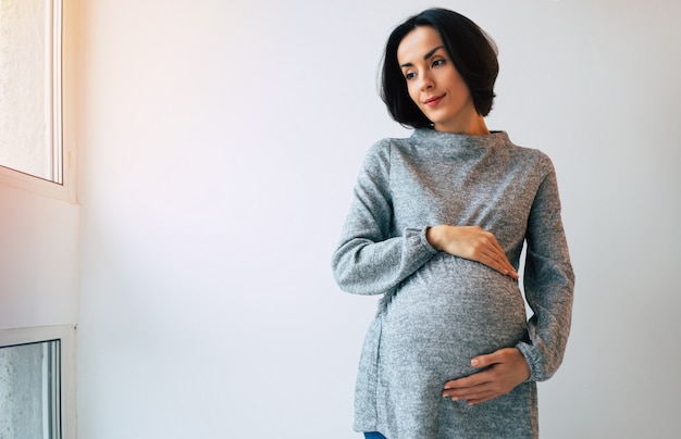 Schitterende zwangere vrouw in een grijze jurk houdt haar buik vast terwijl ze door het raam naar buiten kijkt.