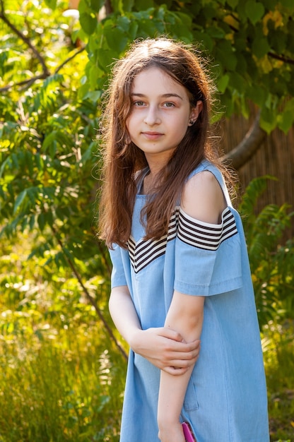 A school-age girl posing against green trees in a dress. Teenager girl in nature.