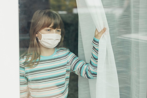 A school-age girl in a protective medical mask looks out the window