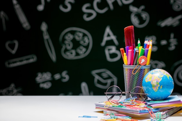 School background with stationery accessories. Books, globe, pencils and various office supplies lying on the desk on a green blackboard background.