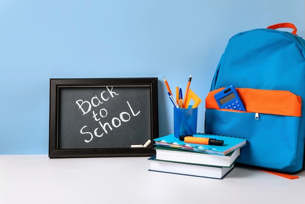 カラフルな学用品と学校に戻る手紙が入った黒板が入った学校のバックパック。青い背景の学用品。バナーデザイン
