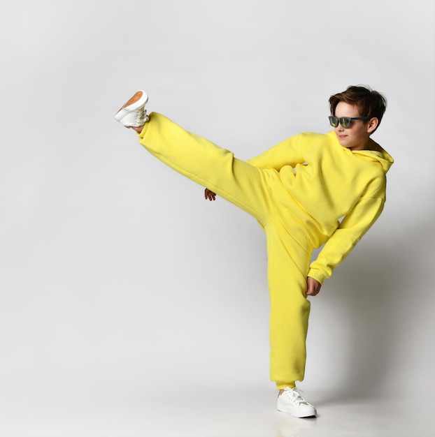 School boy dressed in a stylish bright yellow sports suit and white sneakers posing on a white background Cool child in sunglasses kicks near a free space for text Children's comfortable sportswear