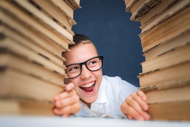 School boy in glasses sitting between two piles of books and look away from camera smiling.