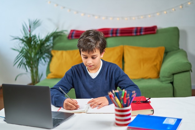 격리 된 집에 앉아 노트북을 사용하고 메모를하는 동안 온라인 수업을하는 학교 소년