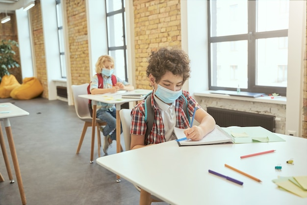 School boy wearing face mask during corona virus and flu outbreak writing in his notebook while