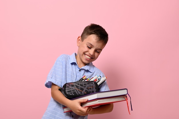 Школьник с пеналом и книгами смеется, позирует на розовом фоне с копией пространства. Концепции возвращения в школу с мимикой и эмоциями