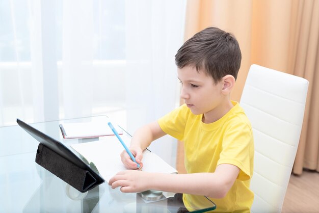Школьник в желтой футболке сидит за столом с цифровым планшетом и делает домашнее задание дома Концепция дистанционного обучения онлайн-образования
