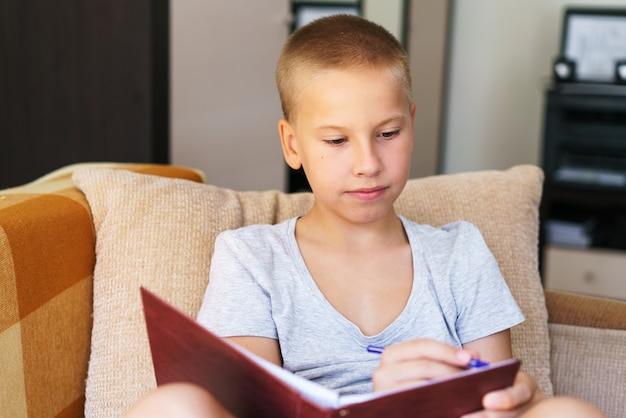 School days boy solves his homework on couch with a notebook in his hands writes answers with a pen ...