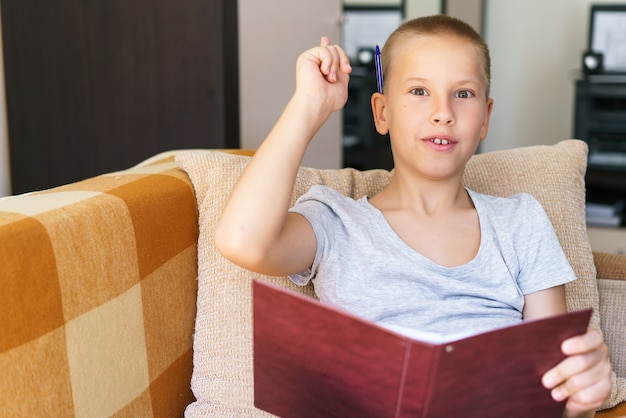 School days boy solves his homework on couch with a notebook in his hands writes answers with a pen ...